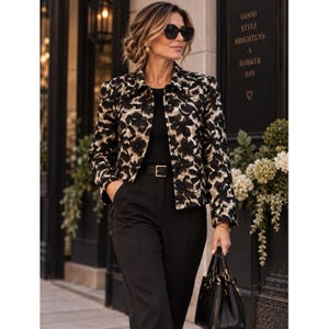 May include: A cropped jacket featuring a black and beige floral pattern, worn over a solid black top and black trousers with a black belt. The jacket has long sleeves, a collared neckline, and a button-front closure. A black handbag is held at the side.