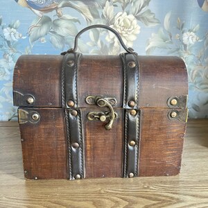 May include: A rectangular wooden chest with a domed lid and a top handle. The dark brown wood features two vertical dark brown leather-like straps secured with metal studs. A central brass-colored metal latch secures the lid to the base.
