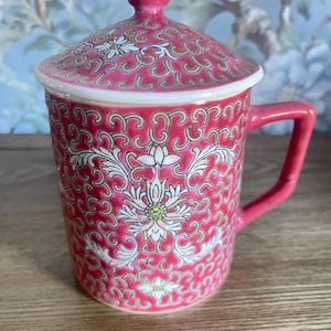 May include: A ceramic mug with a lid featuring a vibrant pink background decorated with intricate white and light green scrolling vine patterns and stylized floral motifs. The mug includes a matching pink handle and a domed lid with a small knob.