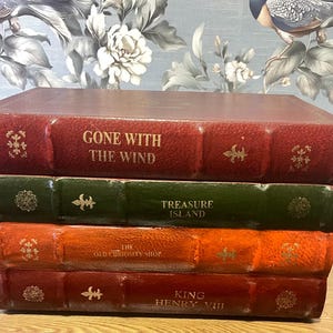 May include: A stack of four hardcover books with textured spines in red, green, orange, and dark red. The titles Gone with the Wind, Treasure Island, The Old Curiosity Shop, and King Henry VIII are printed in gold, accompanied by small gold floral accents.