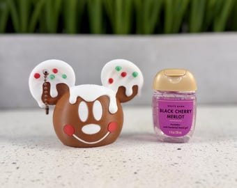 Puede incluir: Un soporte de silicona marrón estilo pan de jengibre con forma de cabeza de ratón con glaseado blanco y chispas rojas y verdes, junto a una pequeña botella transparente de gel antibacteriano White Barn Black Cherry Merlot, 29 ml.