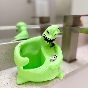 Puede incluir: Una figura hueca de color verde ne&oacute;n brillante con forma de personaje de saco con boca y ojos negros cosidos. La figura presenta una peque&ntilde;a ara&ntilde;a negra en el pecho y sostiene dos dados blancos de seis caras con puntos negros en la mano.