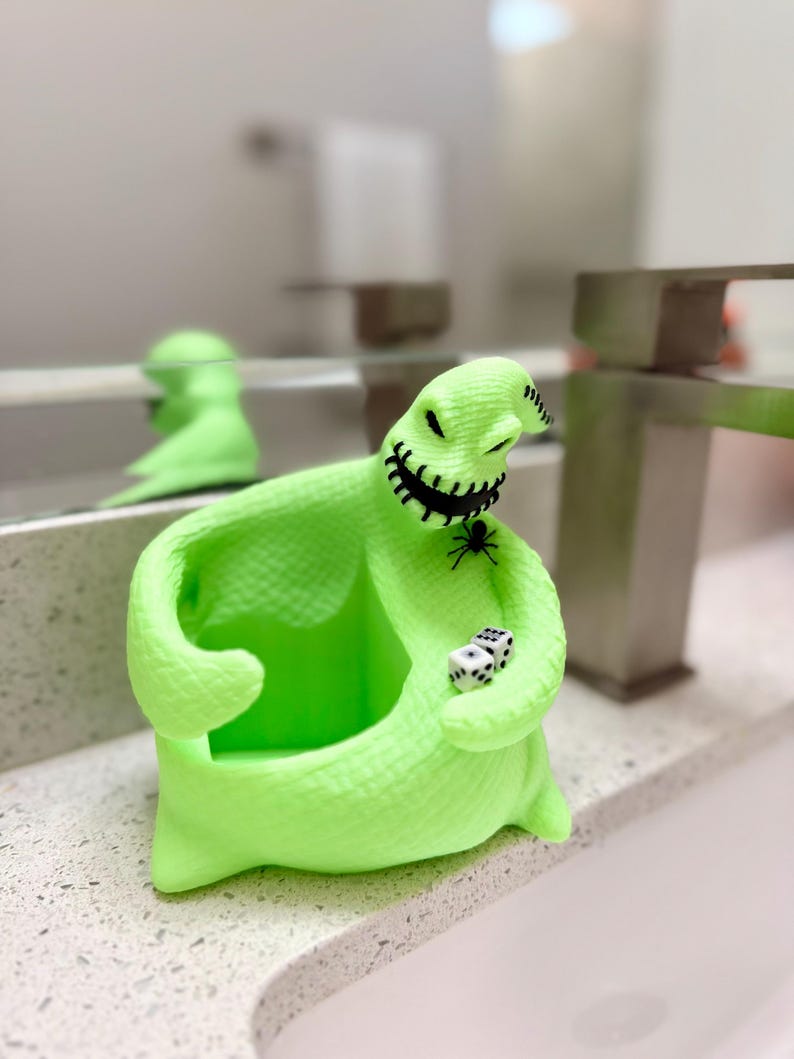 Puede incluir: Una figura hueca de color verde ne&oacute;n brillante con forma de personaje de saco con boca y ojos negros cosidos. La figura presenta una peque&ntilde;a ara&ntilde;a negra en el pecho y sostiene dos dados blancos de seis caras con puntos negros en la mano.