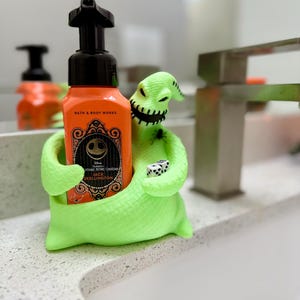 Puede incluir: Un soporte de personaje Oogie Boogie en color verde ne&oacute;n brillante que sostiene una botella de jab&oacute;n naranja de Bath & Body Works con bomba negra. El personaje tiene una boca cosida, una peque&ntilde;a ara&ntilde;a negra en el hombro y dos dados blancos.
