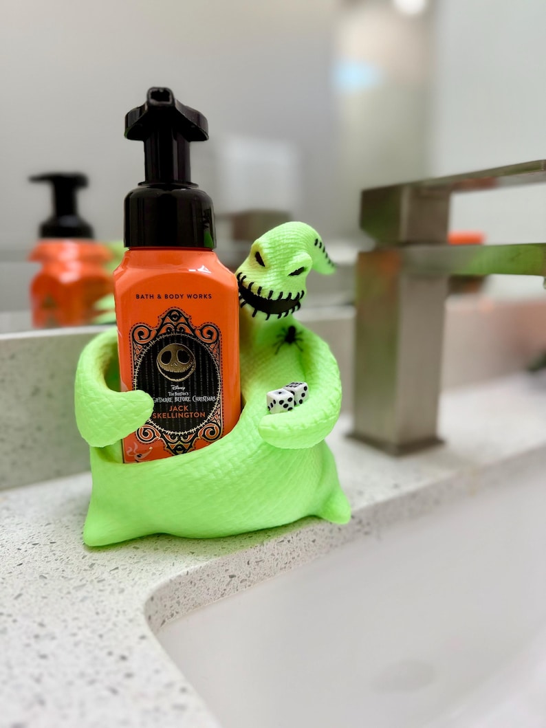 Puede incluir: Un soporte de personaje Oogie Boogie en color verde ne&oacute;n brillante que sostiene una botella de jab&oacute;n naranja de Bath & Body Works con bomba negra. El personaje tiene una boca cosida, una peque&ntilde;a ara&ntilde;a negra en el hombro y dos dados blancos.