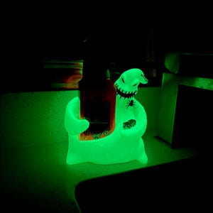 Puede incluir: Una figura verde brillante que se asemeja a un personaje con forma de saco con boca y ojos cosidos, sosteniendo una botella oscura en un brazo y un par de dados en el otro. Un peque&ntilde;o gr&aacute;fico de ara&ntilde;a negra es visible en el pecho.