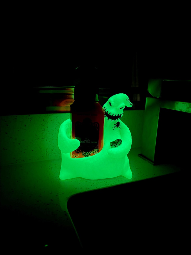 Puede incluir: Una figura verde brillante que se asemeja a un personaje con forma de saco con boca y ojos cosidos, sosteniendo una botella oscura en un brazo y un par de dados en el otro. Un peque&ntilde;o gr&aacute;fico de ara&ntilde;a negra es visible en el pecho.