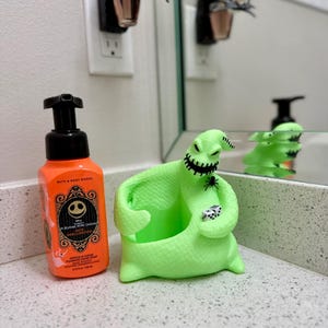 Puede incluir: Una botella de jab&oacute;n de manos en espuma naranja de Bath & Body Works con etiqueta de Jack Skellington junto a un soporte para jab&oacute;n de Oogie Boogie verde lima brillante con una ara&ntilde;a negra y dos dados blancos, y un enchufe de pared de Jack Skellington.