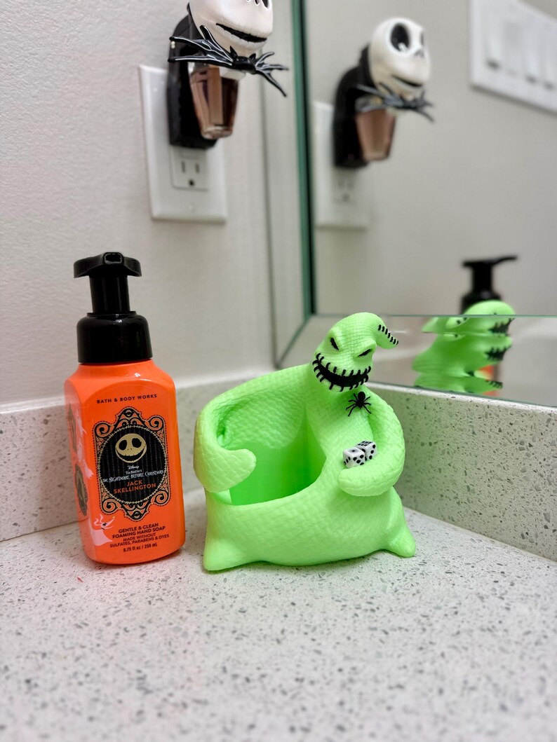 Puede incluir: Una botella de jab&oacute;n de manos en espuma naranja de Bath & Body Works con etiqueta de Jack Skellington junto a un soporte para jab&oacute;n de Oogie Boogie verde lima brillante con una ara&ntilde;a negra y dos dados blancos, y un enchufe de pared de Jack Skellington.