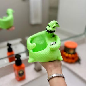 Puede incluir: Un recipiente con forma de saco de color verde ne&oacute;n brillante que representa un personaje con boca cosida, una peque&ntilde;a ara&ntilde;a negra en el pecho y dos dados blancos con puntos negros en la mano. La figura tiene una superficie texturizada y un centro hueco.