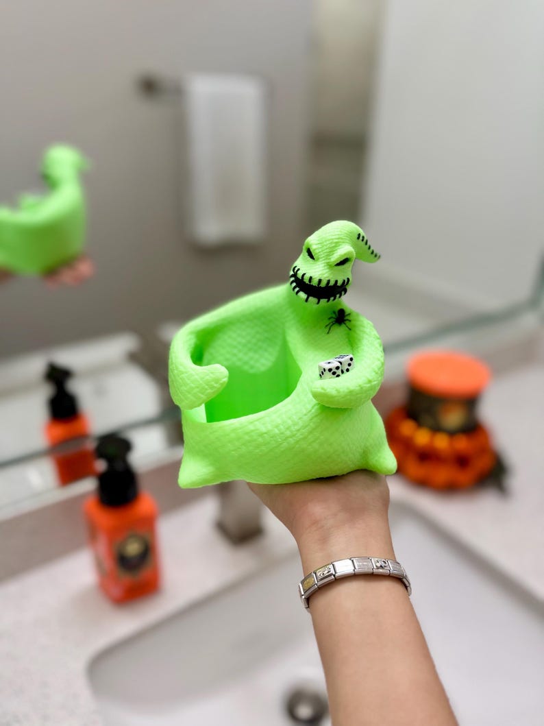 Puede incluir: Un recipiente con forma de saco de color verde ne&oacute;n brillante que representa un personaje con boca cosida, una peque&ntilde;a ara&ntilde;a negra en el pecho y dos dados blancos con puntos negros en la mano. La figura tiene una superficie texturizada y un centro hueco.