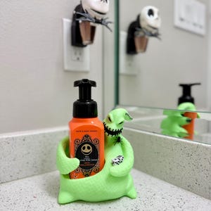 Puede incluir: Un soporte para jab&oacute;n de color verde lima brillante con forma de Oogie Boogie que sostiene una botella de jab&oacute;n en espuma naranja de Bath & Body Works. La botella tiene una etiqueta negra con la cara blanca de Jack Skellington, una ara&ntilde;a negra y dados blancos.
