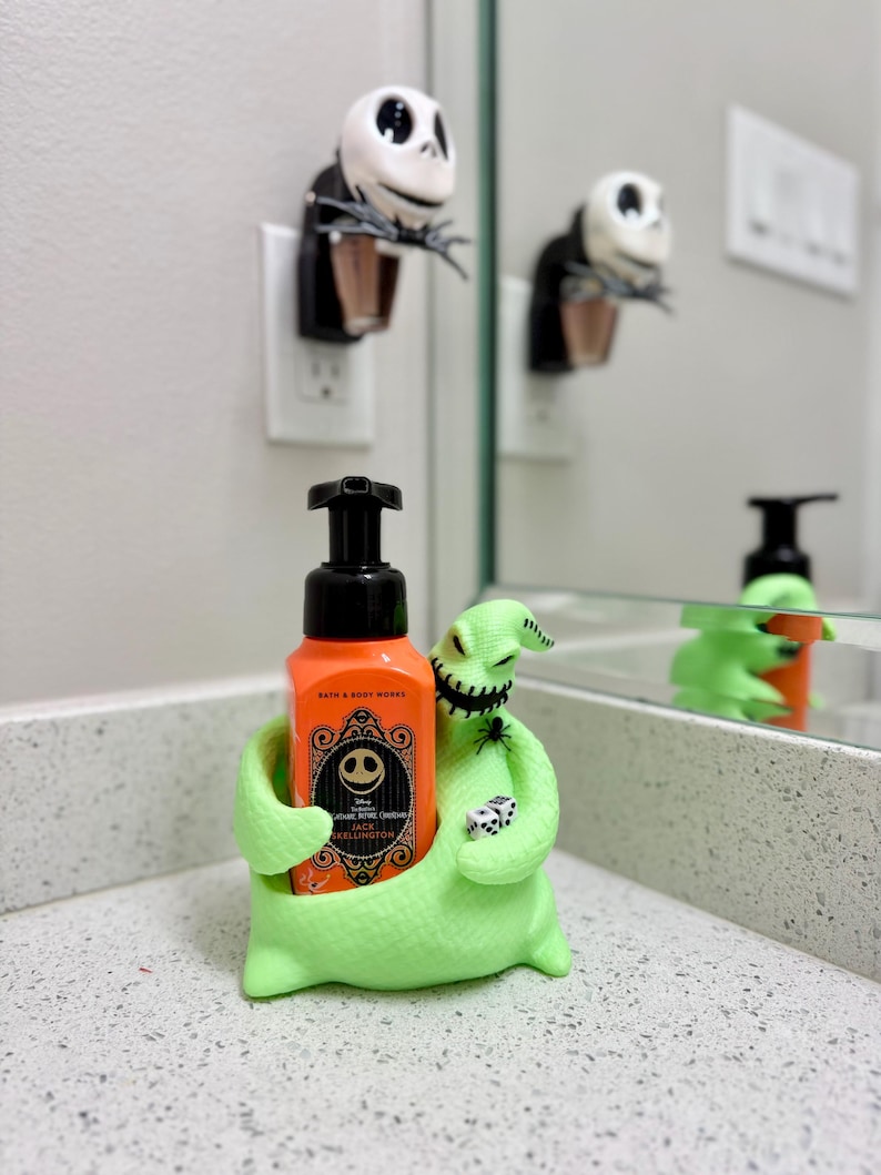 Puede incluir: Un soporte para jab&oacute;n de color verde lima brillante con forma de Oogie Boogie que sostiene una botella de jab&oacute;n en espuma naranja de Bath & Body Works. La botella tiene una etiqueta negra con la cara blanca de Jack Skellington, una ara&ntilde;a negra y dados blancos.