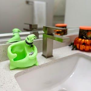 Puede incluir: Una figura texturizada de color verde lima de un personaje con forma de saco, boca cosida, ojos grandes y una peque&ntilde;a ara&ntilde;a negra en el hombro. El personaje sostiene dos peque&ntilde;os dados blancos en sus manos y tiene un centro hueco.