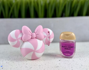 Puede incluir: Un soporte de silicona rosa y blanco con forma de cabeza de ratón con lazo y una pequeña cadena rosa, junto a un frasco transparente de gel antibacteriano para manos White Barn Black Cherry Merlot de 29 ml.
