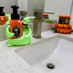 Puede incluir: Un soporte para jab&oacute;n de Oogie Boogie en color verde lima con boca cosida que sostiene un par de dados, conteniendo una botella de jab&oacute;n en espuma naranja de Bath & Body Works con etiqueta de Jack Skellington y bomba negra.
