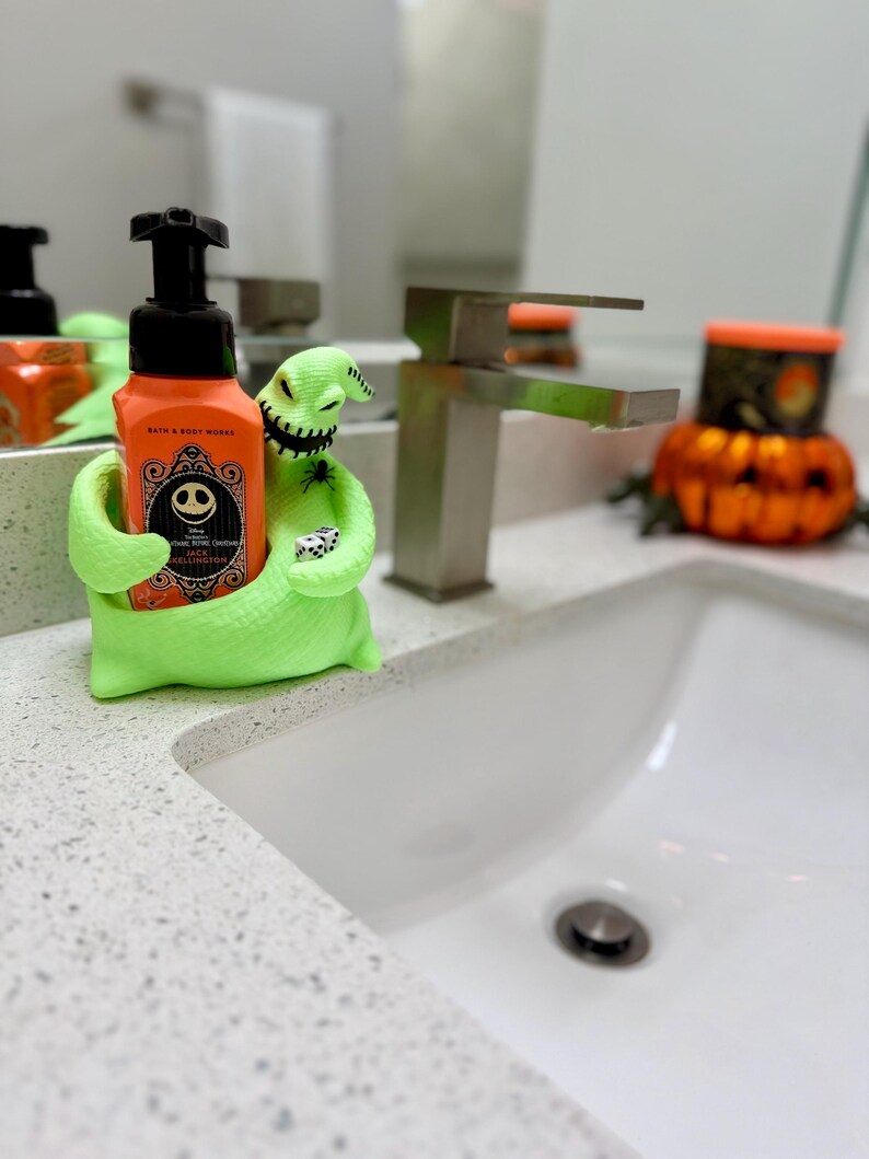 Puede incluir: Un soporte para jab&oacute;n de Oogie Boogie en color verde lima con boca cosida que sostiene un par de dados, conteniendo una botella de jab&oacute;n en espuma naranja de Bath & Body Works con etiqueta de Jack Skellington y bomba negra.