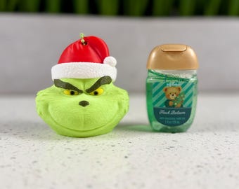 Puede incluir: Un adorno con forma de cabeza de personaje verde que lleva un gorro de Papá Noel rojo y blanco, junto a un pequeño frasco transparente de gel desinfectante verde con tapa dorada y etiqueta con un oso y el texto Fresh Balsam 29 ml.