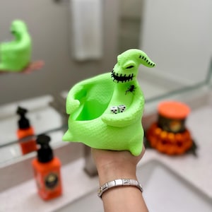 Puede incluir: Un recipiente con forma de personaje de color verde ne&oacute;n brillante y texturizado, con forma de saco y una abertura superior ancha. La figura presenta una boca negra cosida, un peque&ntilde;o gr&aacute;fico de ara&ntilde;a negra y dos dados blancos en las manos.