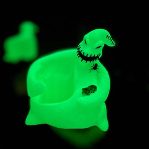 Puede incluir: Una figura que brilla en la oscuridad con forma de personaje de saco, con boca cosida, cabeza puntiaguda y una peque&ntilde;a ara&ntilde;a en el pecho. La figura sostiene dos dados peque&ntilde;os en sus manos, emitiendo una luz verde ne&oacute;n brillante sobre fondo negro.