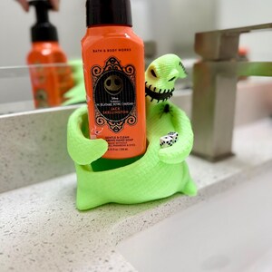 Puede incluir: Un soporte para jab&oacute;n con forma del personaje Oogie Boogie en color verde ne&oacute;n brillante que sostiene una botella de jab&oacute;n de manos en espuma naranja de Bath & Body Works. La etiqueta muestra a Jack Skellington, el texto The Nightmare Before Christmas y 259 ml.