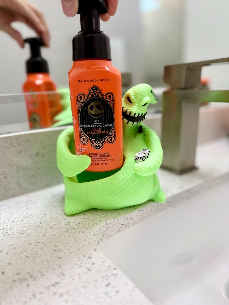 Puede incluir: Un soporte para jab&oacute;n con forma del personaje Oogie Boogie en color verde ne&oacute;n brillante que sostiene una botella de jab&oacute;n de manos en espuma naranja de Bath & Body Works. La etiqueta muestra a Jack Skellington, el texto The Nightmare Before Christmas y 259 ml.