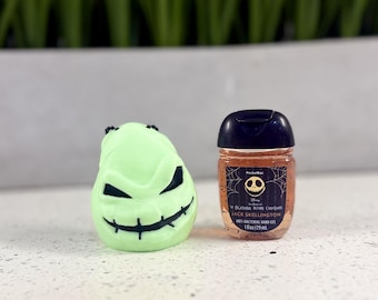 Puede incluir: Una figura de personaje redondeada de color verde claro con boca cosida y ojos afilados, junto a una pequeña botella transparente de gel antibacteriano para manos Jack Skellington con 29 ml de líquido anaranjado.