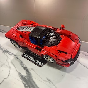 以下が含まれることがあります： 組み立てブロックで作られた赤いフェラーリ・デイトナSP3のモデルカー。車両には黒いルーフ、青い内装シート、シルバーのホイールが備わっており、車体の下にはDAYTONA SP3という白い文字が記された黒いディスプレイプレートが配置されています。