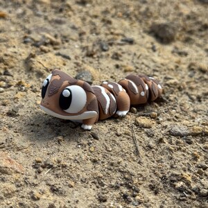 May include: A 3D printed articulated lizard figurine featuring a brown and white patterned body with large, round, white eyes and black pupils. The creature has a segmented, flexible design with visible layer lines, showcasing a smooth, rounded head and a long, tapering body composed of several distinct, connected sections.