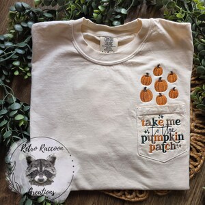 May include: A cream-colored short-sleeve t-shirt featuring an embroidered design of small orange pumpkins above a chest pocket. The pocket displays the embroidered text "take me to the pumpkin patch" in a mix of orange and dark green cursive lettering.