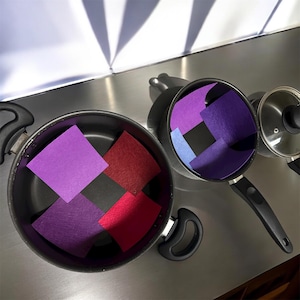 May include: Two black non-stick cooking pots with black handles, each containing square felt pan protectors. The larger pot holds purple and red felt squares, while the smaller pot contains purple and light blue felt squares. A glass lid is visible.