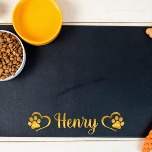 Peut inclure: Un tapis de nourriture pour animaux rectangulaire noir avec le nom Henry en lettres dorées, encadré par deux icônes de pattes en forme de cœur. Le tapis est disposé avec un bol d'eau jaune, un bol blanc de croquettes et deux jouets.