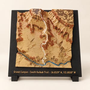 May include: A three-dimensional topographic map of the Grand Canyon South Kaibab Trail mounted on a black frame. The terrain features tan and reddish-brown textures with a white line marking the trail and a blue line representing the river.