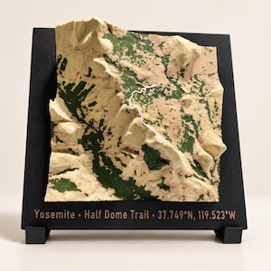 Puede incluir: Un mapa topográfico en 3D del sendero Half Dome de Yosemite montado sobre una base cuadrada negra. El terreno presenta texturas en beige y verde oscuro con una línea blanca que traza el sendero. El texto dice: Yosemite • Half Dome Trail • 37.749°N, 119.523°W.