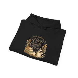 May include: A black hooded sweatshirt featuring a graphic print with the text "Official Member of the Cozy Chapter Club" in cursive script, surrounded by illustrations of a stack of books, a lit candle, and a steaming mug of coffee.