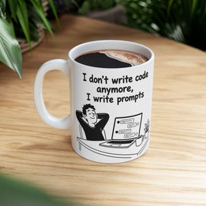 May include: A white ceramic mug featuring a black line drawing of a person sitting at a desk with a laptop, with the text "I don't write code anymore, I write prompts" printed above the illustration. The mug is filled with dark coffee.
