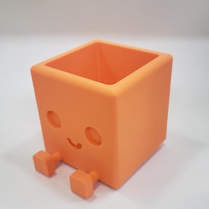May include: A bright orange square-shaped planter with a hollow center. The front face features a recessed design of two circular eyes and a small curved mouth. Two small square feet extend from the bottom front of the container.