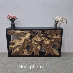 May include: A rectangular console table featuring a black frame enclosing a natural, light brown teak root wood centerpiece. The table surface holds a black vase with pink flowers and a clear vase with white orchids. The text Real photo is visible.