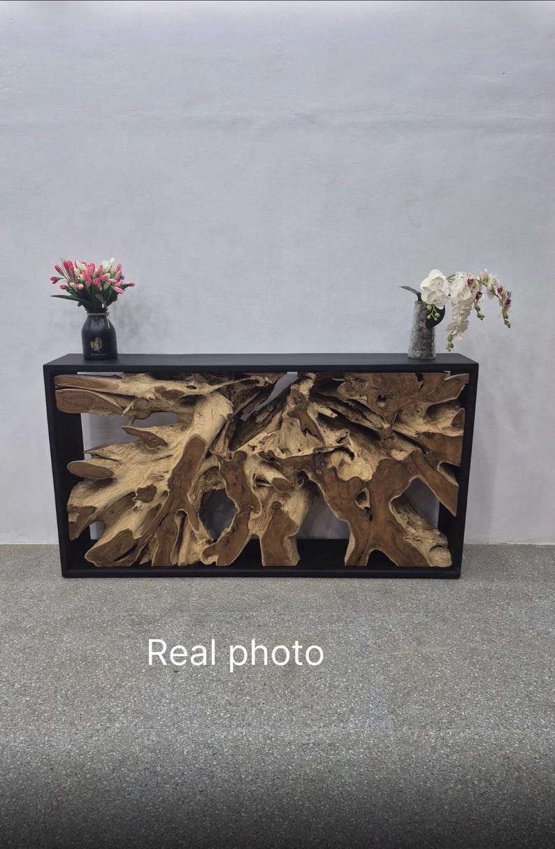 May include: A rectangular console table featuring a black frame enclosing a natural, light brown teak root wood centerpiece. The table surface holds a black vase with pink flowers and a clear vase with white orchids. The text Real photo is visible.