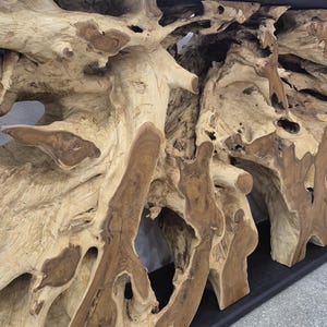 May include: A console table featuring a natural light brown teak root base with intricate, organic wood grain patterns and knots. The base supports a smooth, flat, matte black rectangular top surface, creating a contrast between the raw wood.