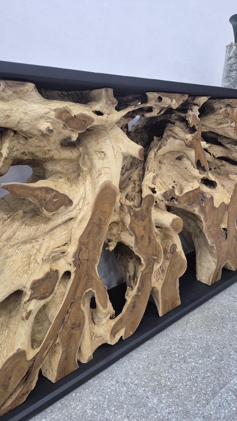 May include: A console table featuring a natural light brown teak root base with intricate, organic wood grain patterns and knots. The base supports a smooth, flat, matte black rectangular top surface, creating a contrast between the raw wood.