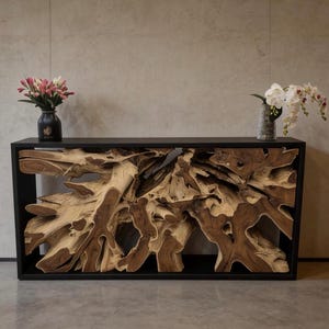 May include: A console table featuring a natural, raw teak root base encased within a sleek, rectangular black metal frame. The top surface is flat and black, supporting a dark vase with pink flowers and a grey vase with white orchids.