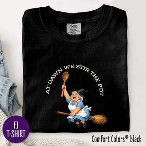 May include: A black crew neck t-shirt featuring a graphic of a smiling character wearing a witch hat and apron, riding a broomstick while holding a wooden spoon. The text AT DAWN WE STIR THE POT is printed in a white arched font above the illustration.