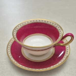 May include: A porcelain teacup and saucer set featuring a vibrant magenta pink band with a delicate white and gold geometric pattern along the rim. The interior of the cup is white, and the handle is accented with gold trim and magenta detailing.
