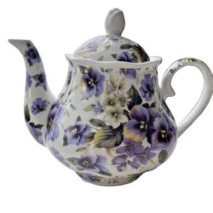 May include: A white porcelain teapot featuring a repeating pattern of purple and yellow pansy flowers with green leaves. The teapot has a curved handle, a long spout, and a removable lid with a small, bud-shaped finial on top.
