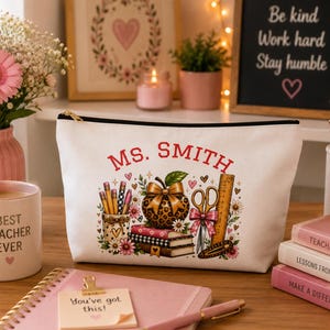 Puede incluir: Un estuche blanco con cremallera que presenta el texto MS. SMITH en letras rojas sobre un gráfico de una manzana con estampado de leopardo, una pila de libros, tijeras con un lazo rosa, una regla y un portalápices, rodeado de flores.