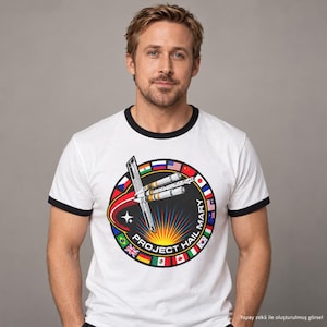 May include: A white ringer t-shirt with black trim on the collar and sleeves. The chest features a circular graphic with a space station, a bright sunburst, and international flags around the border, with the text Project Hail Mary.
