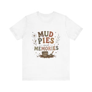 May include: A white short-sleeve t-shirt featuring a graphic print with the text MUD PIES and MEMORIES in brown and orange lettering. The design includes a small brown bucket, a shovel, a daisy, and leafy sprigs surrounded by tiny stars.