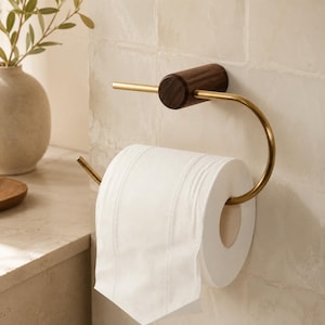 May include: A wall-mounted toilet paper holder featuring a dark brown cylindrical wooden base attached to a curved, brushed gold metal arm. A single roll of white toilet paper is suspended on the metal arm, which extends horizontally.