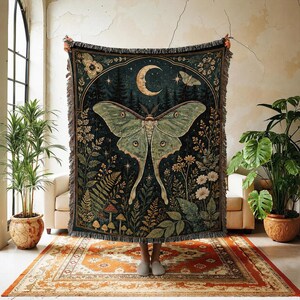 May include: A woven throw blanket featuring a large central luna moth, a crescent moon, and a dark forest background with various plants, ferns, and mushrooms. The design is framed by an archway and includes fringed edges on the top and bottom.